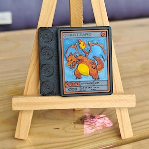 Könnte beinhalten: Eine 3D-gedruckte Charizard-Sammelkarte mit einem feurigen orangefarbenen Drachen vor blauem Hintergrund. Die Karte ist auf einer schwarzen Basis mit kreisförmigen Details befestigt. Präsentiert auf einer kleinen Holzstaffelei.
