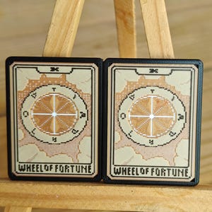 May include: Two "Wheel of Fortune" tarot cards. Each card features a circular design with a wheel and text. The cards have a beige background with brown and black accents. The cards are displayed on a wooden easel.