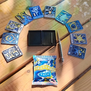 May include: An assortment of fantasy trading cards and accessories displayed on a wooden surface. The cards showcase detailed designs in blue, gold, and white. A package labelled "SPECTRAL PACK" is also present, along with a black case and a metal tool.