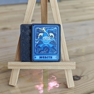 May include: A blue and black card with the word "WRATH" printed on it. The card features a pixelated illustration of a creature with tentacles. The card is displayed on a small wooden easel.