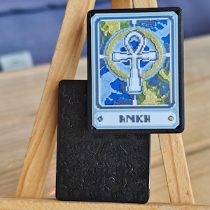 May include: A black and gold Ankh card with a blue and gold design. The card has the word "ANKH" printed on it. The card is displayed on a wooden easel.