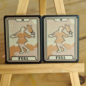 May include: Two tarot cards depicting the Fool. Each card has a black border and a beige background with a pixelated design. The Fool is illustrated in orange and beige, with the word "FOOL" at the base.