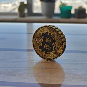 Puede incluir: Una moneda de Bitcoin de color dorado con un símbolo de Bitcoin negro en el centro. La moneda está sobre una superficie de madera.