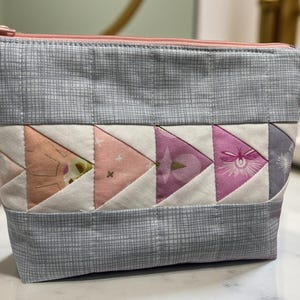 May include: A quilted cosmetic bag with a grey geometric pattern and a pink zip. The bag features a row of colourful triangles in shades of pink, peach, and purple. The bag is designed to hold makeup and other beauty products.