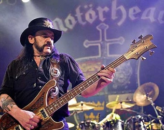 May include: A musician wearing a black shirt and a black top hat plays a brown bass guitar with intricate carvings. The background features the word "Motörhead" in white lettering.