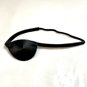 May include: A black leather eye patch with a textured surface. The eye patch is connected to a thin, black elastic band designed to be worn around the head. The eye patch is a classic accessory.