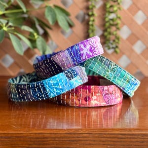 May include: A stack of colorful fabric headbands. The headbands are made of patchwork fabric in shades of blue, purple, green, and red, with white stitching. The headbands are on a wooden surface.