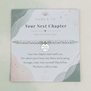 May include: A bracelet with silver beads, light green and pink stones, and a small circular charm that says "You've got this." The bracelet is displayed on a card with the text "Here's to Your Next Chapter."