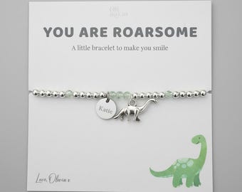 May include: A silver bracelet with a dinosaur charm and a tag engraved with "Katie". The card reads "YOU ARE ROARSOME" and features a green dinosaur illustration. The bracelet is made of small beads.
