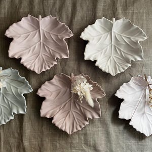 Könnte beinhalten: Fünf blattförmige Keramikschalen in Rosa-, Weiß- und Blaugrüntönen. Einige sind mit kleinen Blumengestecken verziert. Die Schalen sind auf einer neutralfarbenen Stoffoberfläche angeordnet.