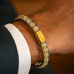 May include: A beaded bracelet featuring light yellow beads and a gold-coloured rectangular clasp. The bracelet is worn on a wrist, with a dark blue suit and white shirt cuff in the background. The bracelet's diameter is approximately 6.35 cm.