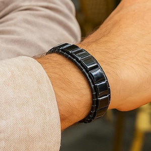 Op de afbeelding: Een zwarte armband met rechthoekige schakels wordt om een pols gedragen. De armband is gemaakt van een donker materiaal en heeft een strak, modern design. De persoon draagt een beige overhemd.