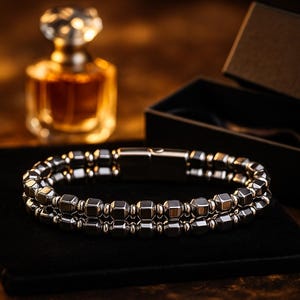 May include: A silver-toned bracelet featuring two strands of geometric beads. The bracelet is presented on a black surface, with a perfume bottle and a black box in the background. The bracelet has a magnetic clasp.
