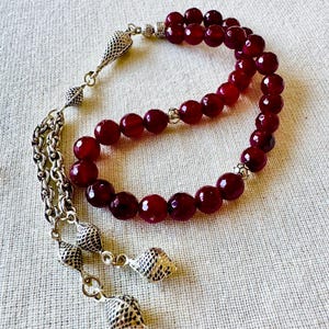 May include: A red agate prayer bead mala with silver accents and tassel. The beads are round and have a smooth, polished finish. The tassel is made of silver chains and has three decorative pendants.