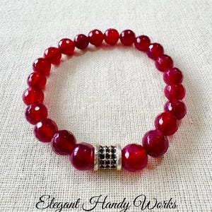 May include: A handmade red beaded bracelet with a silver accent. The bracelet features faceted, round beads and a decorative silver cylinder with black accents. The text "Elegant Handy Works" is visible.