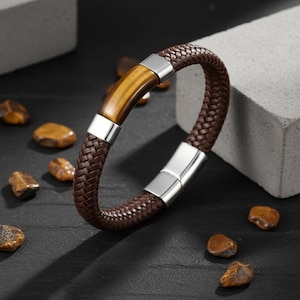 May include: A brown leather bracelet with a woven design. The bracelet features a tiger's eye stone and silver-coloured metal accents. The bracelet is approximately 20 cm in circumference.