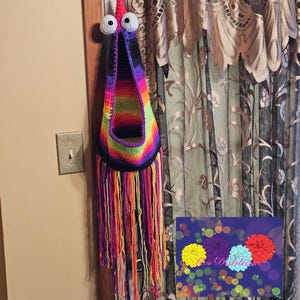 May include: A colourful crocheted hanging basket with a rainbow pattern and black fringe. The basket has two large white googly eyes and a black mouth. The basket is hanging on a wall next to a white light switch.
