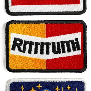 Op de afbeelding: Geborduurde patch met het woord "Rrrttumi" in zwarte letters. De patch heeft een witte rand, een zwarte omtrek en een ontwerp van rode en gele kleuren. De patch is rechthoekig van vorm.