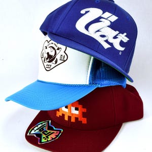 May include: Three baseball caps stacked on top of each other. The top cap is blue with white text that says "U.S.A.T.". The middle cap is white with a blue and green mesh back and a black graphic of a planet with text around it. The bottom cap is maroon with an orange pixelated graphic of a ghost and a holographic sticker on the side.