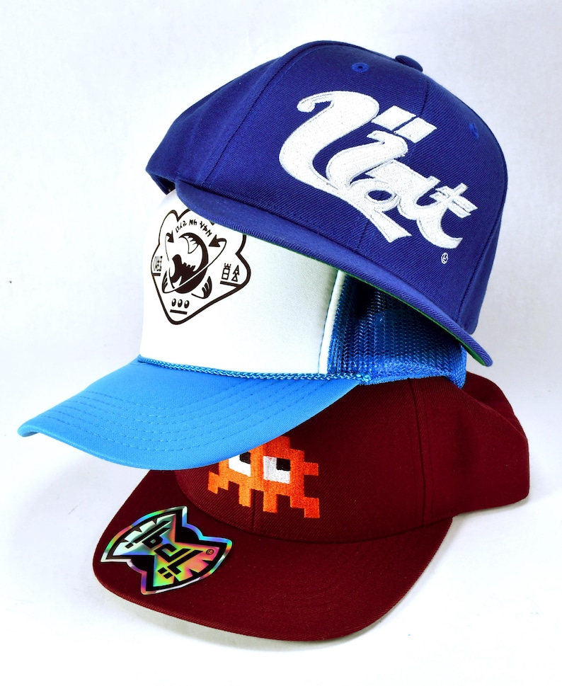May include: Three baseball caps stacked on top of each other. The top cap is blue with white text that says "U.S.A.T.". The middle cap is white with a blue and green mesh back and a black graphic of a planet with text around it. The bottom cap is maroon with an orange pixelated graphic of a ghost and a holographic sticker on the side.