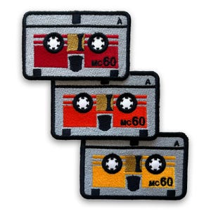 Pode incluir: Três patches bordados em forma de cassetes retro. Cada patch apresenta uma moldura cinzenta, detalhes pretos e um corpo de fita vermelho, laranja ou amarelo. O texto "MC60" é visível em cada patch.