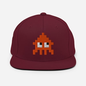 Könnte beinhalten: Burgunderfarbene Baseballkappe mit einem gestickten orangefarbenen, pixeligen Alien-Design mit weißen Augen. Die Kappe hat einen flachen Schirm und eine strukturierte Krone und bietet einen klassischen, lässigen Stil.
