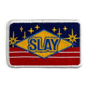 Puede incluir: Un parche rectangular con borde blanco. La sección superior es azul marino con estrellas amarillas. Un diamante amarillo en el centro tiene la palabra "SLAY" en azul. La sección inferior es roja con rayas horizontales blancas.
