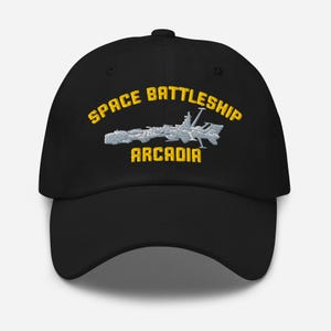 Könnte beinhalten: Schwarze Baseballkappe mit dem gelben Schriftzug "SPACE BATTLESHIP ARCADIA". Unter dem Text befindet sich ein silbernes und weißes Raumschiffdesign. Die Kappe hat einen gebogenen Schirm und eine runde Krone.