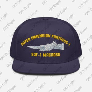 Puede incluir: Una gorra de béisbol azul oscuro con un gráfico bordado de la nave espacial SDF-1 Macross en azul claro y blanco. El texto "SUPER DIMENSION FORTRESS-1" y "SDF-1 MACROSS" está bordado en amarillo sobre y debajo del gráfico de la nave.