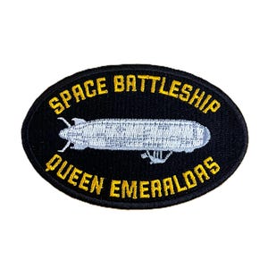 Puede incluir: Parche ovalado negro con el texto amarillo "SPACE BATTLESHIP QUEEN EMERALDAS". Un diseño de nave espacial plateada y blanca está en el centro. El parche está bordado.