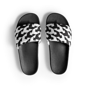 May include: A pair of black slide sandals. The top strap features a repeating pattern of black arrows on a white background. The footbed is black with a textured surface, and the sandals are set against a white background.