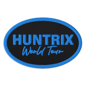 May include: An embroidered patch with a black oval center and a blue border. The patch features the word "HUNTRIX" in blue, above the words "World Tour" in a smaller, stylized font.