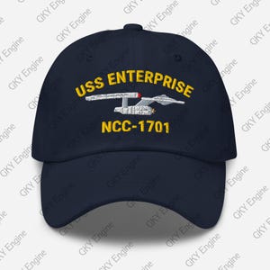 Könnte beinhalten: Eine marineblaue Baseballkappe mit einer gestickten Grafik des Raumschiffs USS Enterprise in Weiß und Grau. Der Text USS ENTERPRISE ist in Gelb über dem Schiff gestickt, und NCC-1701 ist in Gelb unter der Schiffsgrafik gestickt.