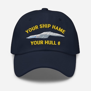 May include: A navy blue baseball cap featuring yellow embroidered text that reads YOUR SHIP HAME and YOUR HULL #. Between the two lines of text is an embroidered graphic of a grey starship with blue and red accents on its side.