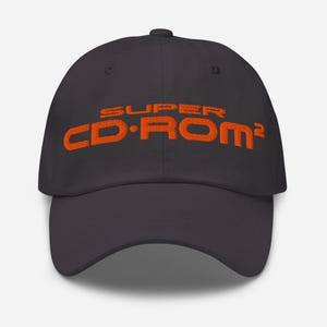 Peut inclure: Une casquette de baseball noire avec le texte SUPER CD-ROM² brodé en fil orange vif sur le panneau avant. La casquette présente une calotte structurée, une visière incurvée et un bouton sur le dessus, offrant un design sobre et épuré.