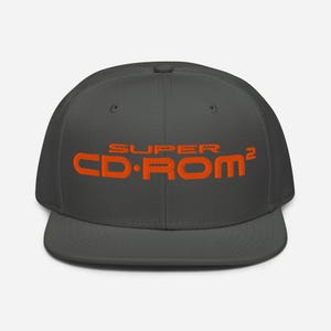 Peut inclure: Une casquette de baseball gris foncé avec le texte SUPER CD-ROM² brodé en fil orange vif sur le panneau avant. La casquette présente une calotte structurée, une visière plate et un bouton sur le dessus, offrant un design simple et épuré.