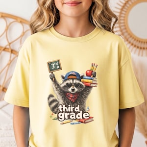 May include: A pale yellow t-shirt featuring a graphic of a raccoon wearing sunglasses, a baseball cap, and a red bandana. The raccoon holds a small chalkboard labeled 3rd, a stack of books, and an apple. The text third grade is printed below.