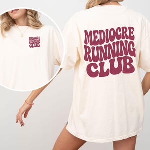 May include: A cream-colored short-sleeve t-shirt featuring the text "MEDIOCRE RUNNING CLUB" in a large, bold, dark red wavy font on the back. A smaller version of the same text and font is printed on the upper left chest area of the shirt.