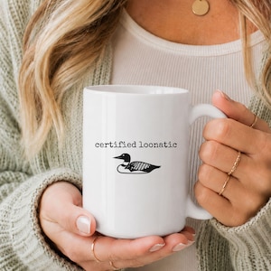May include: A white ceramic mug featuring a black graphic of a loon bird centered on the front. Above the bird, the text "certified loonatic" is printed in a simple, black, typewriter-style font. The mug has a smooth finish and a curved handle.