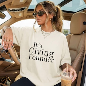 May include: Cream-colored oversized t-shirt with the text "It's Giving Founder" in black. The shirt is worn by a person in a car, accessorized with jewelry and sunglasses. A beverage in a clear cup is held in the hand.