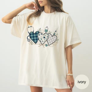 May include: An ivory t-shirt featuring a graphic print of three heart shapes. The hearts are decorated with blue plaid, floral, and grey gingham patterns. A stethoscope, an IV bag, and a syringe are integrated into the heart design.