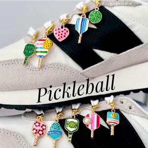 May include: A pair of white sneakers with black accents and colorful pickleball paddle charms attached to the laces. The charms feature different designs, including a heart, a paddle with the words "Let's Play", and a paddle with the word "Pickleball".