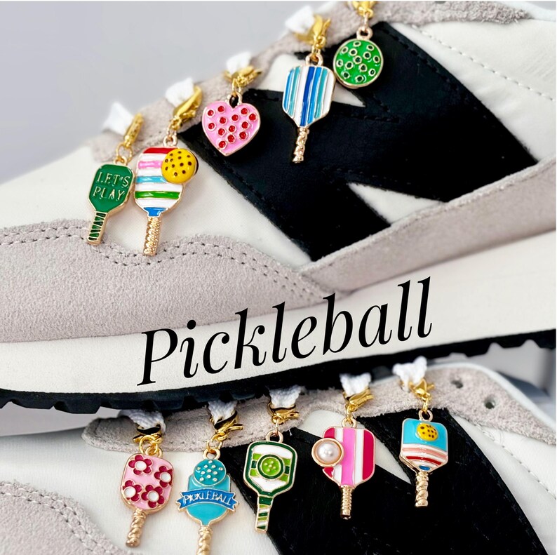 May include: A pair of white sneakers with black accents and colorful pickleball paddle charms attached to the laces. The charms feature different designs, including a heart, a paddle with the words "Let's Play", and a paddle with the word "Pickleball".