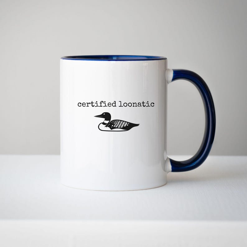May include: A white ceramic mug with a dark blue interior and handle. The front features a black graphic illustration of a loon bird positioned below the black text that reads "certified loonatic" in a simple, typewriter-style font.