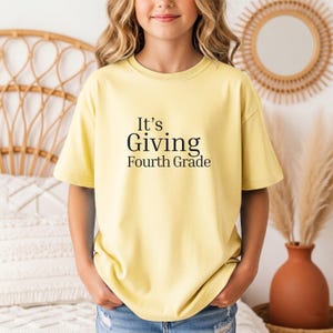 May include: A pale yellow short-sleeved t-shirt featuring the black text It's Giving Fourth Grade printed on the front. The shirt has a crew neckline and a relaxed fit, constructed from a solid-colored fabric with no additional patterns or designs.