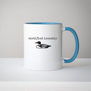 May include: A white ceramic mug with a bright blue interior and handle. The front features a black graphic illustration of a loon bird positioned below the black text that reads "certified loonatic" in a simple, typewriter-style font.