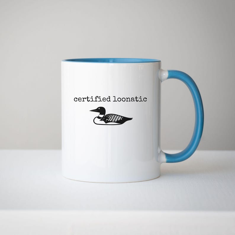 May include: A white ceramic mug with a bright blue interior and handle. The front features a black graphic illustration of a loon bird positioned below the black text that reads "certified loonatic" in a simple, typewriter-style font.