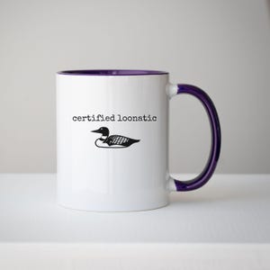 May include: A white ceramic mug with a purple interior and a purple handle. The front of the mug features a black graphic illustration of a loon bird positioned below the black text that reads, "certified loonatic" in a typewriter font.