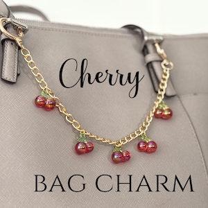 Puede incluir: Un charm de bolso de cadena dorada con charms de cereza rojos y verdes. El charm está unido a un bolso de cuero marrón. El texto "Cherry Bag Charm" está escrito debajo del charm.
