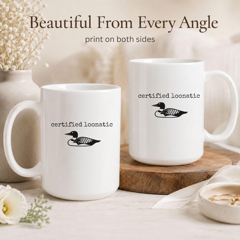 May include: Two white ceramic mugs featuring a black graphic of a loon bird and the text "certified loonatic" printed on both sides. The mugs have a standard handle and cylindrical shape, displaying the design clearly on the front and side.